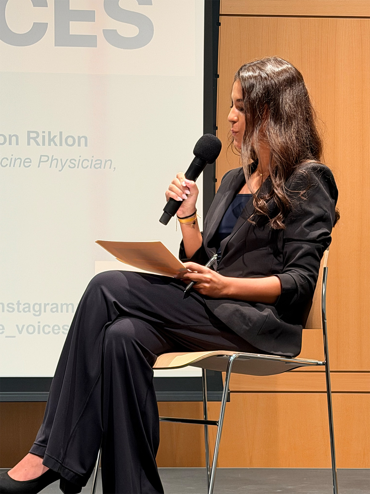 Leyla Tore moderating from her chair, microphone and notes in hand.